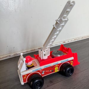 Könnte beinhalten: Ein Vintage-Feuerwehrauto-Spielzeug von Fisher-Price. Der rot-weiße Truck hat eine weiße Leiter, schwarze Räder und eine Feuerwehrfigur. Der Text "FISHER PRICE FIRE ENGINE" ist auf der Seite aufgedruckt.