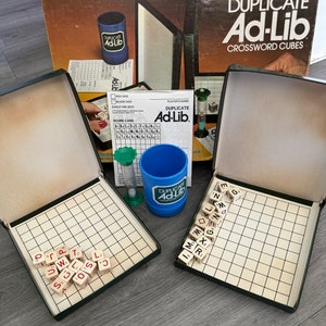 May include: A vintage board game called "Duplicate Ad-Lib Crossword Cubes". The game includes a green and white hourglass, two blue cups with the game's name, and two game boards with wooden letter cubes.
