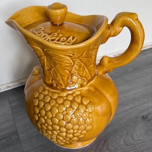 May include: A glossy mustard-yellow ceramic cookie jar shaped like a pitcher with a lid. The surface features embossed grape clusters and leaf patterns. The word Cookies is embossed on the lid, which has a small knob handle on top.