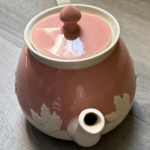 Op de afbeelding: Roze keramische theepot met een witte rand en deksel. De theepot heeft een witte handgreep en tuit. Witte bladeren zijn in reli&euml;f op de zijkant aangebracht. Het deksel heeft een kleine, ronde knop. Een charmante toevoeging aan elke theeservies.