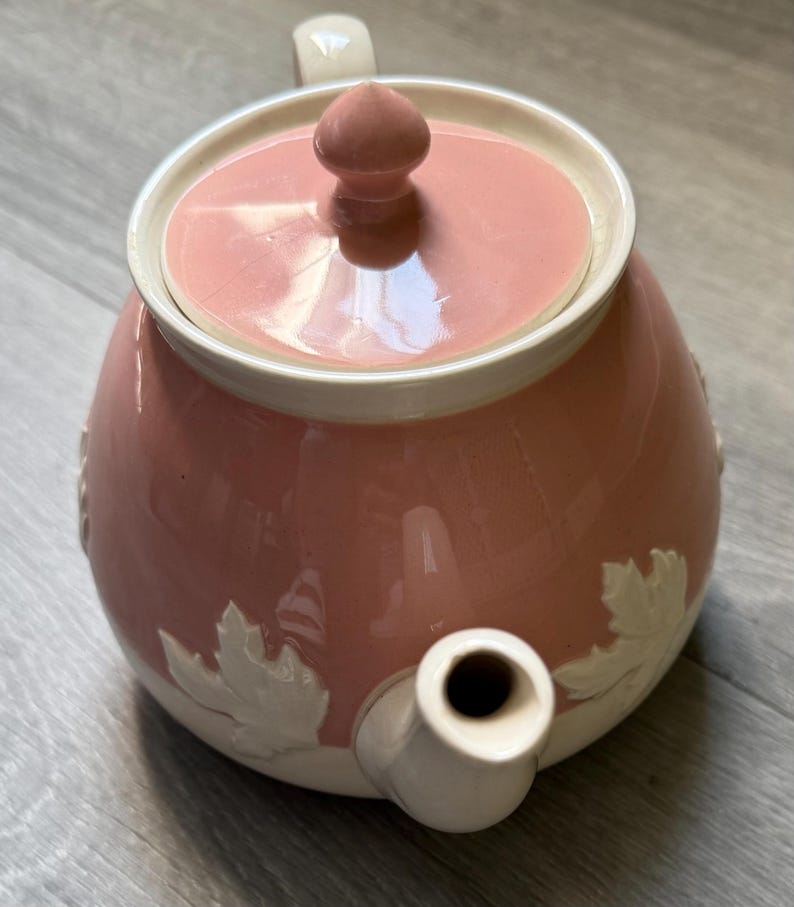 Op de afbeelding: Roze keramische theepot met een witte rand en deksel. De theepot heeft een witte handgreep en tuit. Witte bladeren zijn in reli&euml;f op de zijkant aangebracht. Het deksel heeft een kleine, ronde knop. Een charmante toevoeging aan elke theeservies.