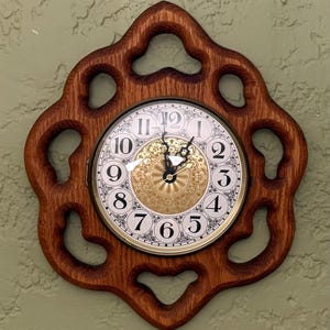 May include: A wall clock featuring a circular face with black Arabic numerals and a gold-toned center with an intricate floral pattern. The clock is encased in a dark brown wooden frame with a scalloped, open-work design and black hands.