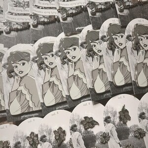 以下が含まれることがあります： 「All along there was an invisible string tying you to me」というテキストと、髪に花をつけた女性が描かれた銀色のエナメルピンセット。ピンは、「primrose」と「vol. 8」のテキストが書かれた白いカードに飾られています。