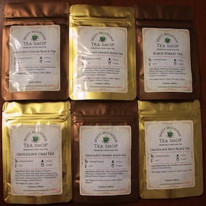 May include: Six gold foil pouches of Misty Mountain Tea Shop loose leaf tea. The pouches are labeled with the tea flavor: Swiss Hot Cocoa Black Tea, Chocolate Orange Black Tea, Black Forest Tea, Chocolate Chai Tea, Chocolate Caramel Black Tea, and Chocolate Mint Black Tea. Each pouch has a label with the ingredients, brewing instructions, and origin of the tea.