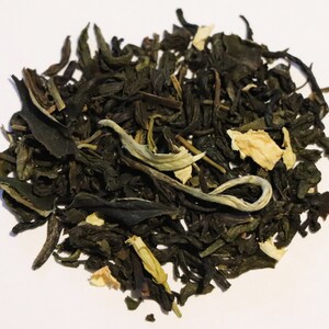 May include: A close-up of a pile of loose leaf green tea with white flower petals. The tea is a blend of green tea leaves and white jasmine flowers.