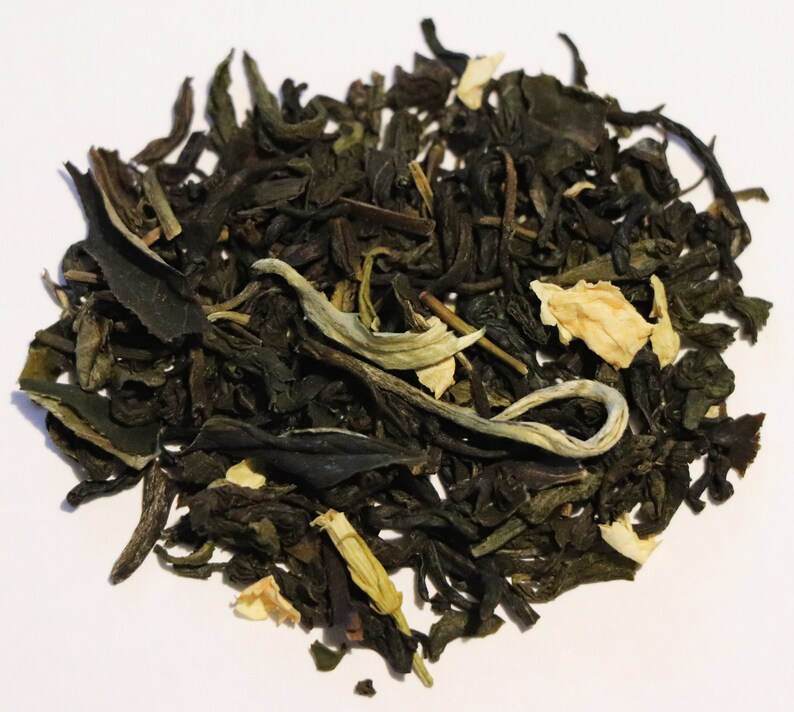 May include: A close-up of a pile of loose leaf green tea with white flower petals. The tea is a blend of green tea leaves and white jasmine flowers.