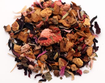 May include: A close-up of a pile of loose leaf tea. The tea blend includes dried pieces of fruit, herbs, and flowers in shades of red, brown, and green. The tea is scattered on a white surface.