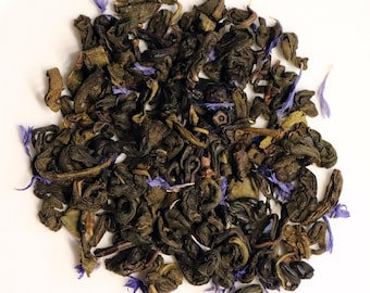 May include: A close-up of a pile of loose leaf green tea with dried cornflower petals. The tea is a dark green color and the petals are a light blue.