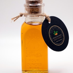 May include: A small glass bottle filled with honey, with a cork stopper and a black and gold label that reads "Misty Mountain Tea Shop, Pure Local Honey, Orange Blossom Raw Honey, Net Wt 2 oz".