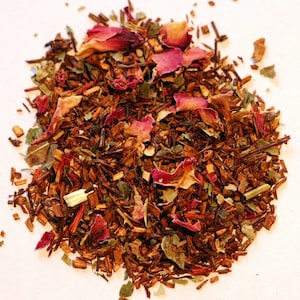 May include: A close-up of a pile of loose leaf tea with red rose petals, brown tea leaves, and green herbs.