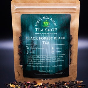 May include: A brown paper pouch with a label that reads "Misty Mountain Tea Shop Premium Loose Leaf Tea Black Forest Black Tea". The label has a green background with a teacup and leaves design. The pouch is open and loose leaf black tea is spilling out.