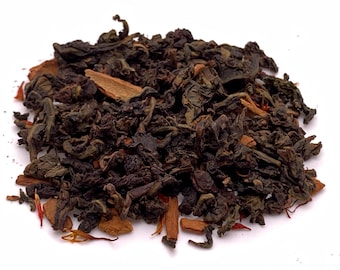 May include: A close-up of loose leaf tea with cinnamon sticks and other spices. The tea leaves are dark green and brown, with pieces of cinnamon and other reddish-brown spices mixed in. The tea is on a white background.