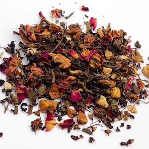 May include: A pile of loose leaf tea with a variety of colors and textures. The tea blend includes dried green leaves, reddish-pink rose petals, and small, tan-colored fruit pieces. The tea is scattered on a white surface.
