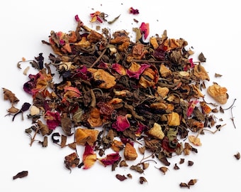 May include: A pile of loose leaf tea with a variety of colors and textures. The tea blend includes dried green leaves, reddish-pink rose petals, and small, tan-colored fruit pieces. The tea is scattered on a white surface.