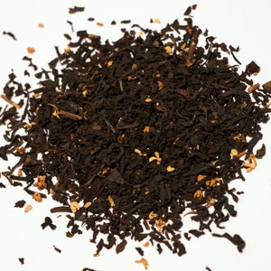 May include: A close-up of a pile of loose leaf black tea with small orange pieces. The tea is scattered on a white surface.