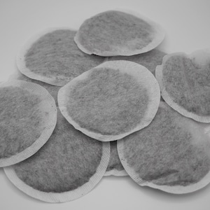 May include: A pile of round, gray tea bags with white edges, arranged on a white surface. The tea bags have a soft, textured appearance, suggesting a loose-leaf tea blend. The image is a close-up shot.