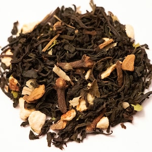 May include: Close-up of loose-leaf tea blend with dark tea leaves, cloves, and pieces of dried fruit. The tea blend has a variety of textures and colors, including dark brown, tan, and light brown. The tea is scattered on a white surface.