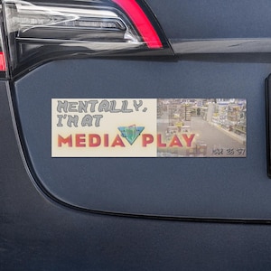 May include: A rectangular bumper sticker on a dark blue car. The sticker reads "Mentally, I'm at Media Play" in a colorful, retro font. A small image of a store is to the right of the text.
