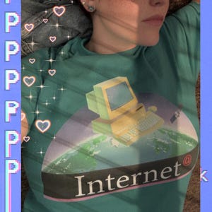 Könnte beinhalten: Türkisfarbenes T-Shirt mit einer Retro-Computergrafik über einem Globus und dem Wort "Internet" darunter. Das Shirt hat eine Vintage-Ästhetik. Das Bild ist mit einem blauen Rahmen mit dem Buchstaben "P" auf der linken Seite versehen.