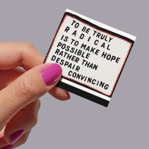 May include: A small, rectangular card with the text "TO BE TRULY RADICAL IS TO MAKE HOPE POSSIBLE RATHER THAN DESPAIR CONVINCING" is held by a person. The card has a white background with a black border and red accents. The person's nails are painted pink.
