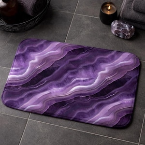 Può includere: Un tappetino da bagno rettangolare con un design a forma di agata viola. Il tappetino presenta fasce a spirale di viola intenso, lavanda e bianco, creando un effetto marmorizzato. Il tappetino è posizionato su un pavimento piastrellato grigio scuro.