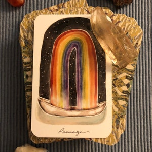 Könnte beinhalten: Eine Aquarell-Tarotkarte mit einem Regenbogen, der sich über einem Boot wölbt, vor einem sternenklaren Nachthimmel. Das Wort "Passage" steht unten. Ein klarer Quarzkristall und dekorative Kartenränder umgeben die Karte.