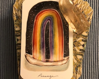 以下が含まれることがあります： 星空を背景に、ボートの上に虹が描かれた水彩画のタロットカード。下部には「Passage」の文字。透明な水晶と装飾的なカードの縁がカードを囲んでいます。