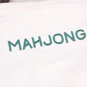 May include: Cream-colored tote bag with the word "MAHJONG" embroidered in green thread. The bag has a simple design with reinforced stitching along the seams and straps, suitable for carrying items.