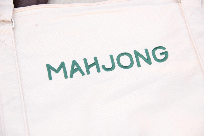 May include: Cream-colored tote bag with the word "MAHJONG" embroidered in green thread. The bag has a simple design with reinforced stitching along the seams and straps, suitable for carrying items.