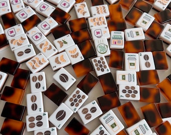 May include: A collection of rectangular mahjong tiles with a white face and a brown tortoiseshell pattern on the back. The tiles feature various graphics including coffee beans, croissants, cafe buildings, and directional markers like North, South, East, and West.