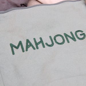 May include: Close-up of a light green canvas tote bag with the word "MAHJONG" embroidered in green thread. The bag has adjustable straps and a zippered top. The bag is designed for carrying mahjong tiles.