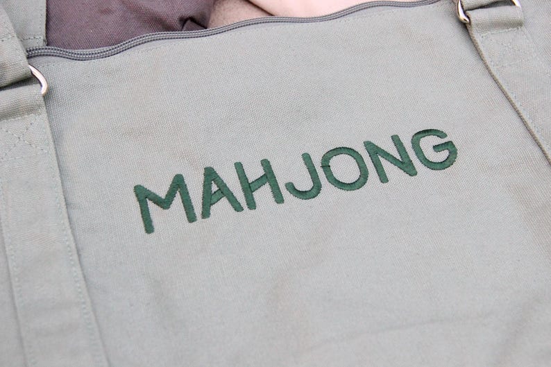 May include: Close-up of a light green canvas tote bag with the word "MAHJONG" embroidered in green thread. The bag has adjustable straps and a zippered top. The bag is designed for carrying mahjong tiles.