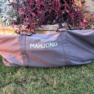 May include: A gray canvas Mahjong bag with white lettering, featuring two handles and a zipper closure. The bag is lying on green grass, with a backdrop of purple and green foliage.