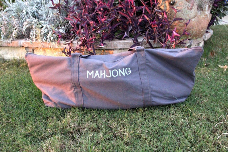 May include: A gray canvas Mahjong bag with white lettering, featuring two handles and a zipper closure. The bag is lying on green grass, with a backdrop of purple and green foliage.