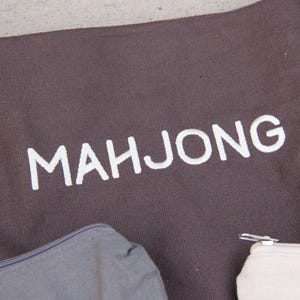May include: A brown tote bag with the word "MAHJONG" printed in white. The bag has two handles and a shoulder strap. A gray zippered pouch and a cream-colored pouch are visible at the bottom.