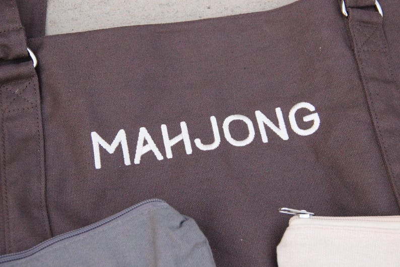 May include: A brown tote bag with the word "MAHJONG" printed in white. The bag has two handles and a shoulder strap. A gray zippered pouch and a cream-colored pouch are visible at the bottom.