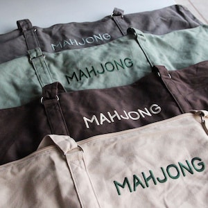 May include: A stack of canvas tote bags in shades of gray, green, brown, and beige. Each bag has a matching strap and the word "MAHJONG" embroidered across the front. The bags are designed for carrying items.