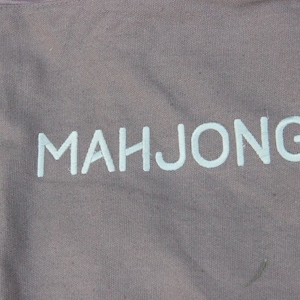 May include: Gray canvas tote bag with the word "MAHJONG" printed in light blue. The bag has a shoulder strap with a metal ring. The fabric has a textured appearance.