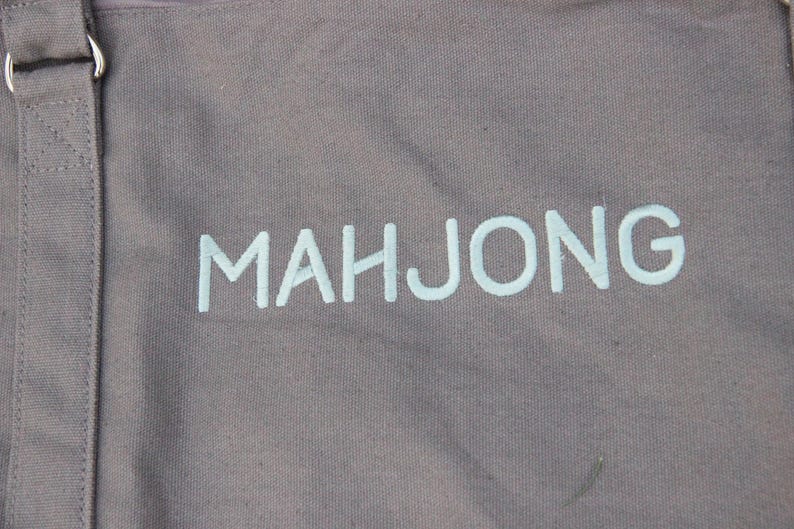 May include: Gray canvas tote bag with the word "MAHJONG" printed in light blue. The bag has a shoulder strap with a metal ring. The fabric has a textured appearance.