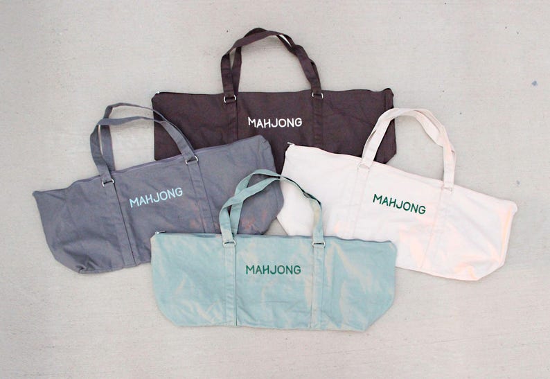 May include: Five tote bags in various colors: dark brown, gray, white, and light blue. Each bag has a rectangular shape with handles and the word "MAHJONG" printed on the front in a contrasting color.