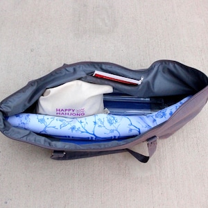 May include: Open tote bag with a gray exterior and a blue floral patterned interior. Inside are a white bag with "HAPPY MAHJONG" printed in purple, a red notepad, and other items. The bag is open on a concrete surface.