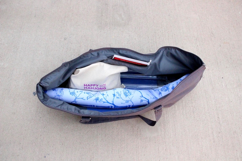 May include: Open tote bag with a gray exterior and a blue floral patterned interior. Inside are a white bag with "HAPPY MAHJONG" printed in purple, a red notepad, and other items. The bag is open on a concrete surface.