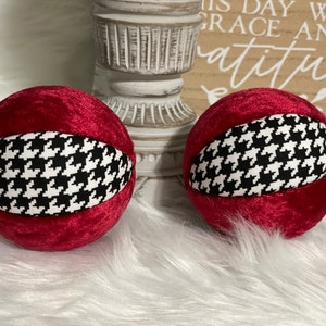 May include: Two red velvet ornaments with a black and white houndstooth pattern. The ornaments are round and have a smooth texture.