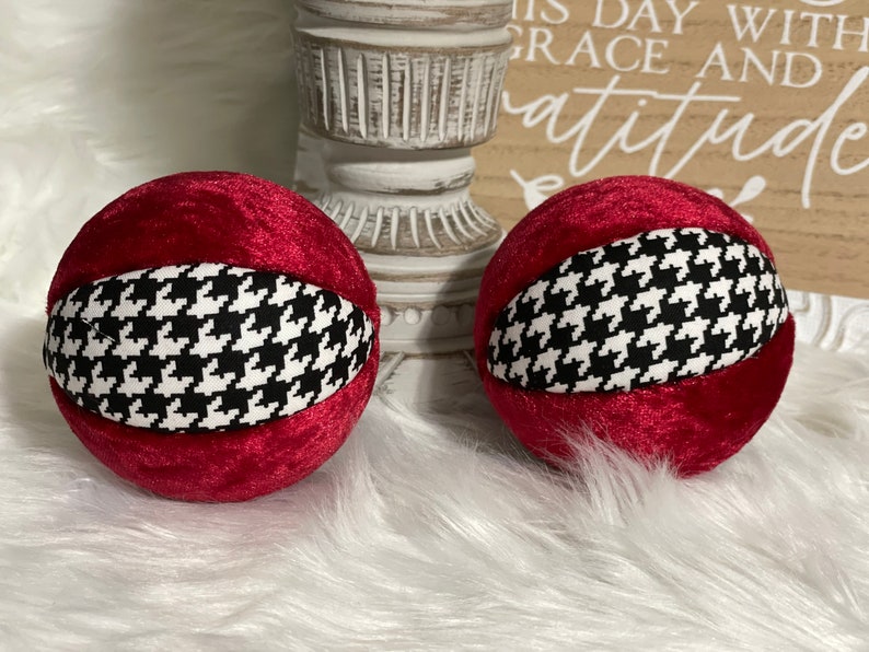 May include: Two red velvet ornaments with a black and white houndstooth pattern. The ornaments are round and have a smooth texture.