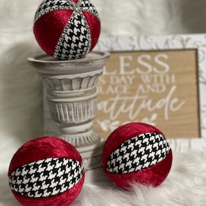 May include: Three red velvet Christmas ornaments with a black and white houndstooth pattern. The ornaments are sitting on a white pedestal and a wooden sign with the text "Bless each day with grace and attitude".