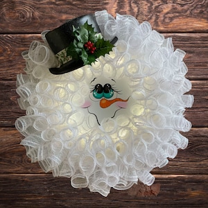 May include: A white mesh wreath with a snowman face and a black top hat with green holly and red berries.