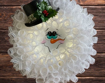 Op de afbeelding: Een witte mesh-krans met een sneeuwpopgezicht en een zwarte hoed met groene hulst en rode bessen.