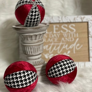 May include: Three red velvet Christmas ornaments with a black and white houndstooth pattern. The ornaments are sitting on a white pedestal and a wooden sign with the text "Bless each day with grace and attitude".