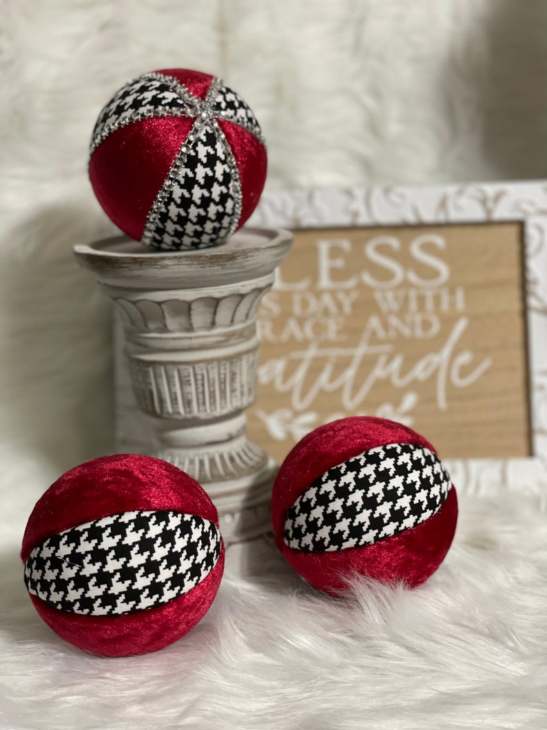 May include: Three red velvet Christmas ornaments with a black and white houndstooth pattern. The ornaments are sitting on a white pedestal and a wooden sign with the text "Bless each day with grace and attitude".
