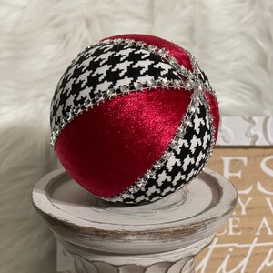 May include: A red and black Christmas ornament with a houndstooth pattern and silver rhinestones. The ornament is sitting on a white pedestal.